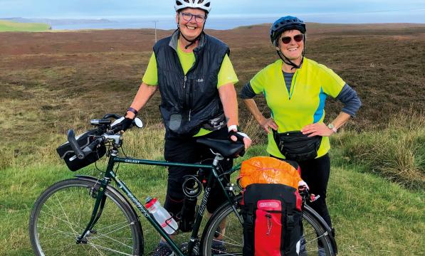 Two women stand behind a Dawes Galaxy touring bike that's loaded with panniers. They are both wearing helmets and high vis tops and smiling broadly at the camera. In the background is a coastline covered in heather.