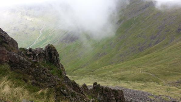 A view of Black Sail Pass, part of Wainwright’s Coast to Coast trail, showing a pass through mountains. A gravel path runs along the bottom of the pass, with grassy mountains rising up on either side. Mist is rolling in.