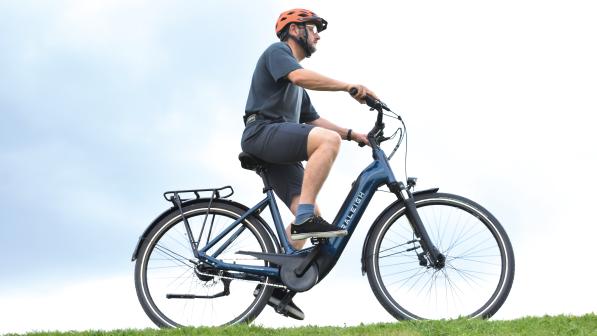 A man wearing a bright orange helmet rides an e-bike on a green field against a blue-sky backdrop. He is wearing a casual t-shirt, trainers and shorts. 