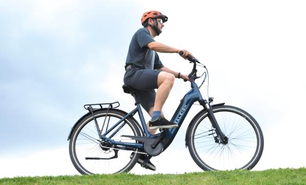 A man wearing a bright orange helmet rides an e-bike on a green field against a blue-sky backdrop. He is wearing a casual t-shirt, trainers and shorts. 