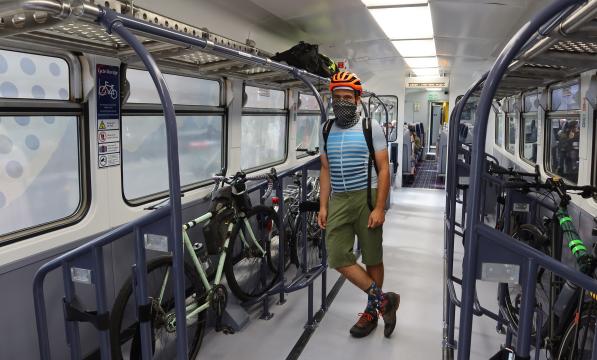 A man stands wearing a helmet. He‘s leaning against the pole in a train bicycle carriage in front of his gravel bike
