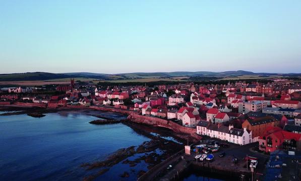 A birds eye view of a Scottish coastal town. A port is in the foreground, red-roofed houses and buildings in the background