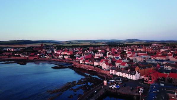 A birds eye view of a Scottish coastal town. A port is in the foreground, red-roofed houses and buildings in the background