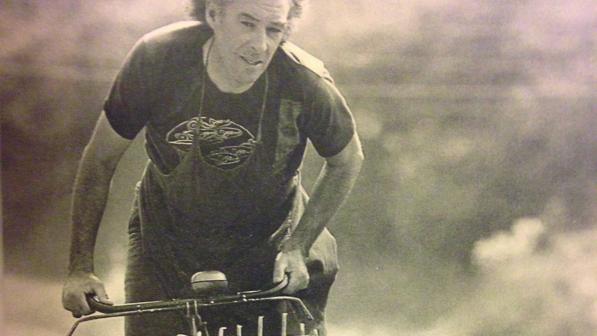 A sepia photo of a man in T-shirt and apron riding a sit-up-and-beg bike with a wicker basket on the front