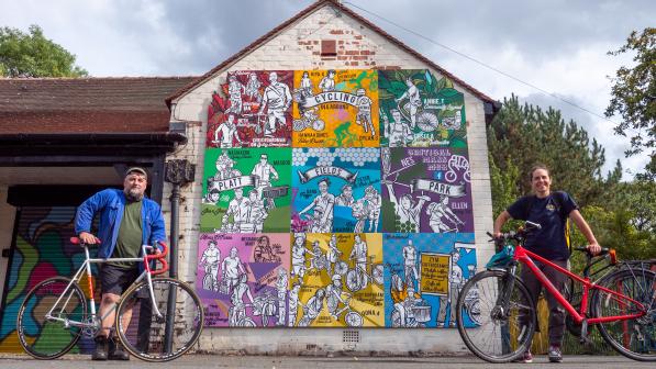 man and woman stand with bike each in front of colourful mural on gable end of old building in park