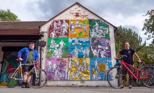man and woman stand with bike each in front of colourful mural on gable end of old building in park