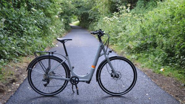 A grey step-through e-cycle stands in the middle of a cycle trail between a wooded area