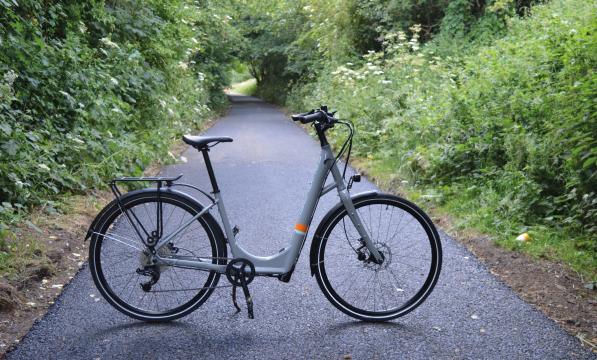 A grey step-through e-cycle stands in the middle of a cycle trail between a wooded area