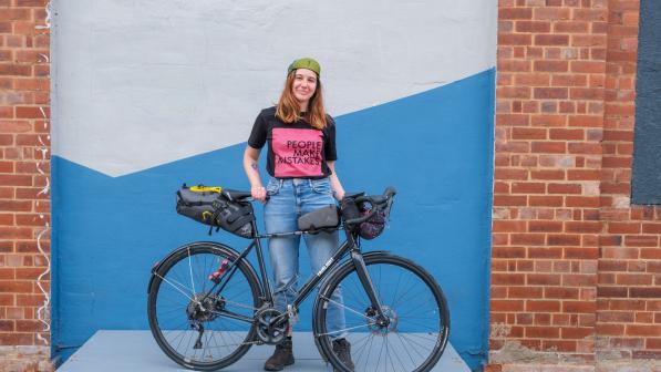 Adna stands smiling with her black bike. She is wearing a cap, black and pink slogan t-shirt and jeans. 