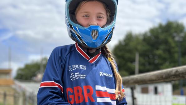 Amelie stands with her BMX bike. She is wearing a BMX helmet, gloves, blue British Cycling jersey and green trousers. 