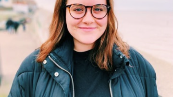 Amy stands with the beach behind her. She is wearing glasses and a dark jacket. 