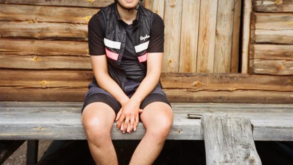 Anna sits on a wooden veranda. She wears a helmet, black top, gilet, shorts and trainers. 
