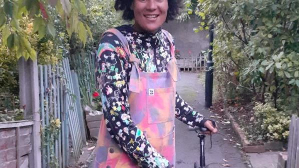 Belinda stands to the left of her bike on a path. She is wearing colourful dungarees and a black long-sleeved top. 