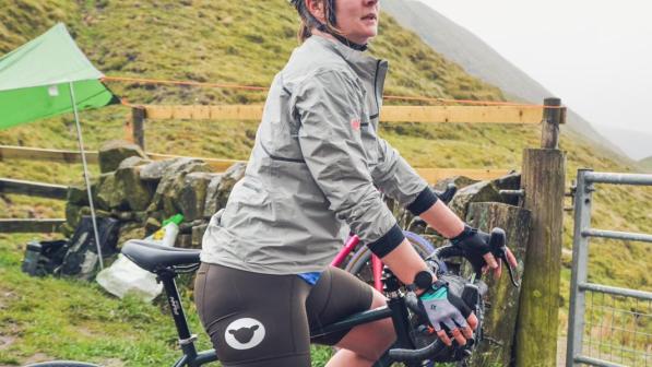Charlotte stands side-on to the camera with her bike in a mountainous landscape. She is wearing a white helmet, grey jacket and brown shorts. 