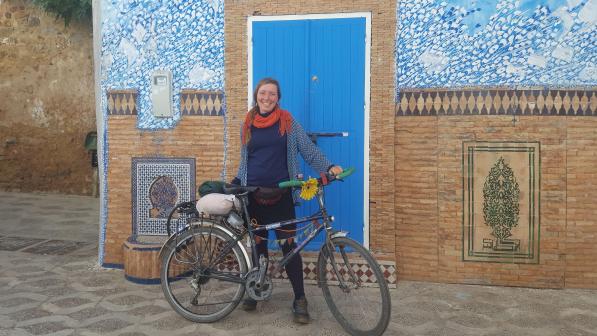Eilidh smiles with her bike during one of her cycle adventures overseas. She is wearing a blue top and red scarf. Behind her is a colourful, historic building.  Her bike has a big yellow flower attached to the handlebars.