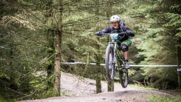 Emma is smiling as her bike does a jump on a mountain biking trail. She is wearing a helmet, dark coloured top and green leggings.