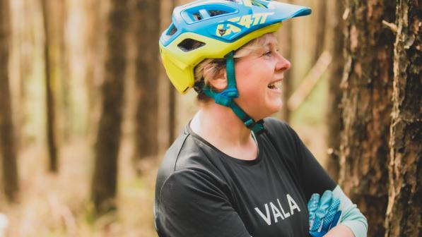 Emma smiles with her arms folded. She is wearing a blue and yellow helmet, black top and blue gloves. There are trees in the background.