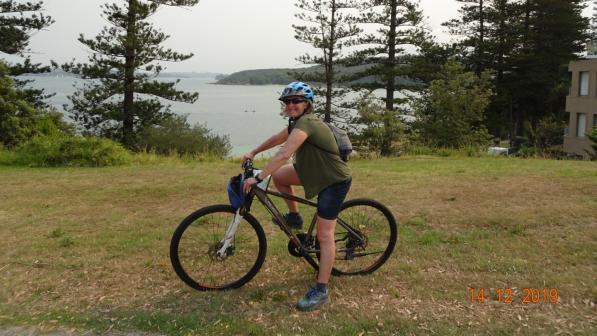 Helen smiling on her bike. She wears a blue helmet, green top and dark shorts. There is a lake behind her.