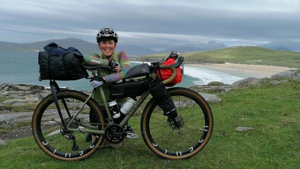 Iona kneels behind her green bike which is set up with bikepacking bags. She wears a helmet, green jersey, gloves and black shorts. A beach is in the background. 