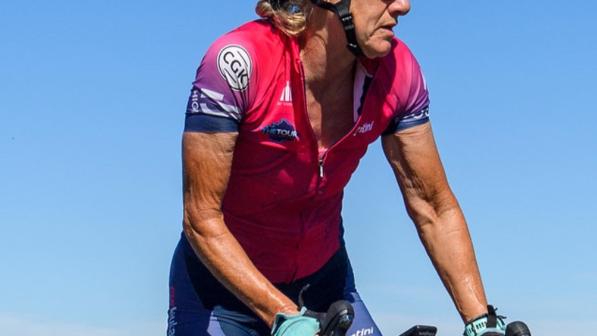 Jackie riding her blue bike in the countryside. She is wearing a pink jersey, blue shorts, black helmet and sunglasses.