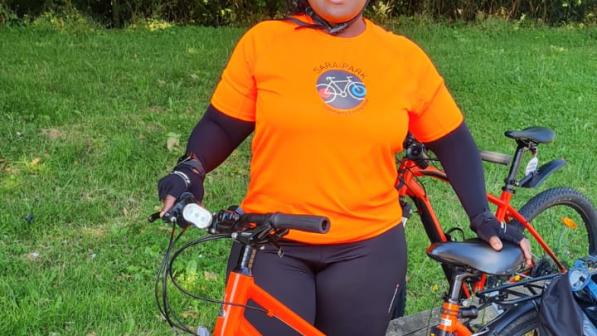 Joscelyne smiling with her red bike. She is wearing an orange Sara Park CCC t-shirt, black leggings and helmet. 