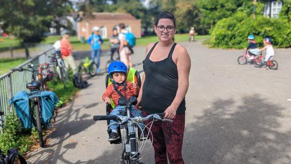 Kat stands to the right of her bike in a park. A small child is in a child’s seat at the back of the bike. Kat is wearing glasses, a black top and red patterned leggings. 