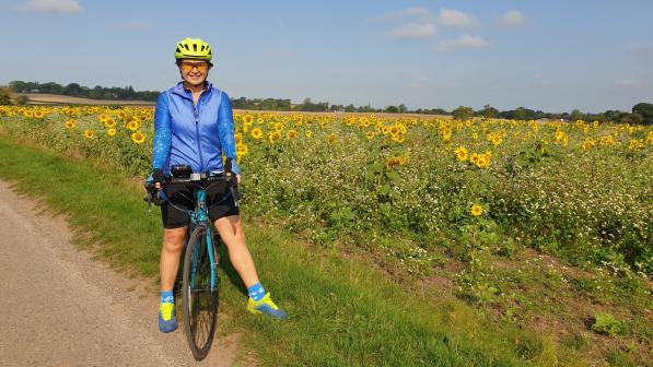 Marina stands next to a sunflower field with her blue bike. She wears sunglasses, a yellow helmet, blue jacket and black shorts. 