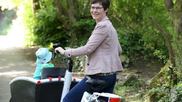 Saskia smiles at the camera as she stands with her cargo bike, transporting her children. She is wearing glasses, a beige jacket and jeans. 
