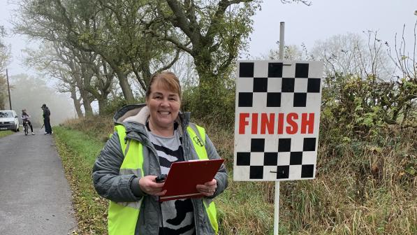 Sharon smiles to camera, holding a clipboard and stopwatch. She is standing at the side of the road at the finish line. She is wearing a grey winter coat and hi-vis vest.