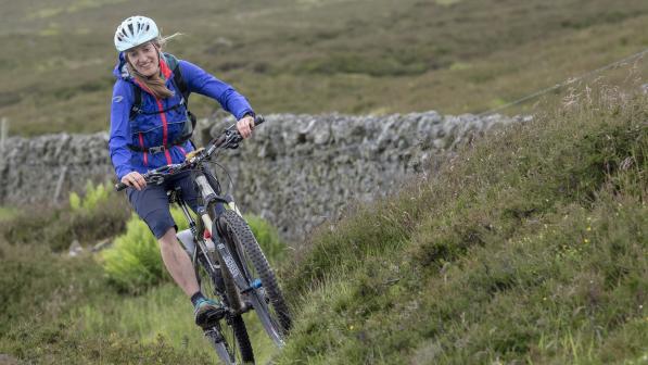 Sophie mountain biking on the Great North Trail. She is wearing a white helmet, blue jacket and grey shorts. 