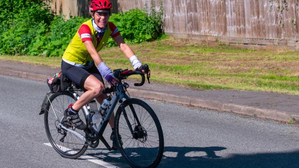 Yasmin smiling as she rides a white Trek bike on road. She is wearing a red helmet, yellow gilet over a red jersey and black cycling shorts. 