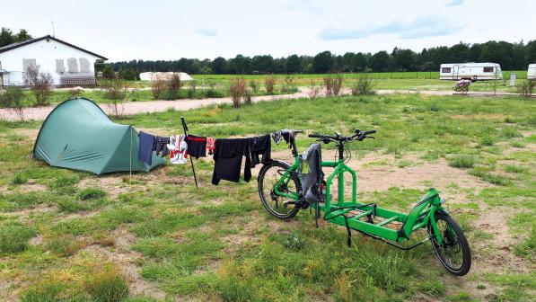 A tent sits on a grassy campsite, with a washing line of cycling clothes hung between it and a bright green cargo bike