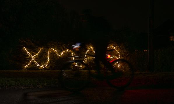 The word 'XMAS' is written out in fairy lights in the background. In front, a cyclist rides past in a blur