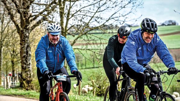 Three men cycle along a road with green fields in the background. 