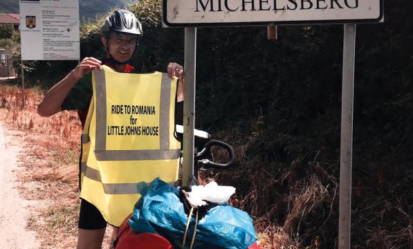 A man stands in front of a road sign that says 'Cisnadioara Michelsberg'. He is holding up a high-vis tabard that bears the words 'Ride to Romania for Little John's House'. He is wearing a helmet and his bicycle leans next to the road sign.