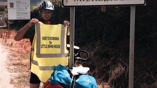 A man stands in front of a road sign that says 'Cisnadioara Michelsberg'. He is holding up a high-vis tabard that bears the words 'Ride to Romania for Little John's House'. He is wearing a helmet and his bicycle leans next to the road sign.