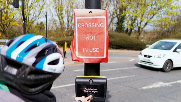 A woman goes to press the button for a pedestrian crossing but the lights have a cover over them which reads 'Crossing not in use'. A car approaches in the background