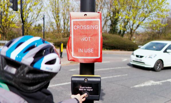 A woman goes to press the button for a pedestrian crossing but the lights have a cover over them which reads 'Crossing not in use'. A car approaches in the background