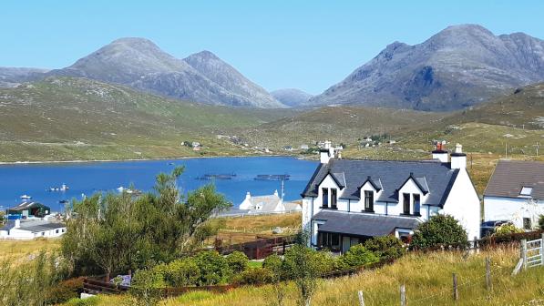 A white cottage sits in a green field in front of a lake and mountains