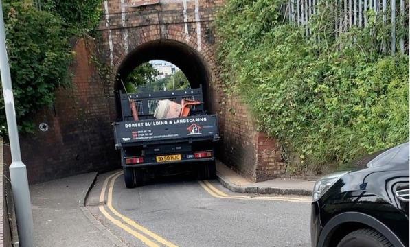 A flat-bed truck is trying to squeeze through a very narrow railway bridge