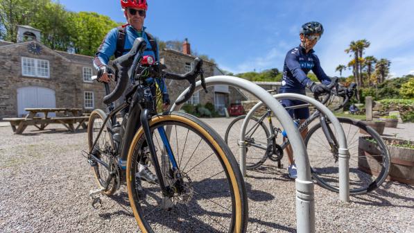 Two men wearing helmets push their bikes into mental bike racks on gravel
