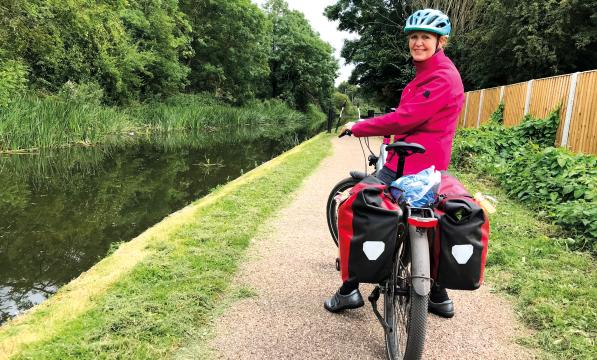 A woman sat on a bicycle laden with full rear panniers turns to look at the camera. She has paused on a canal towpath