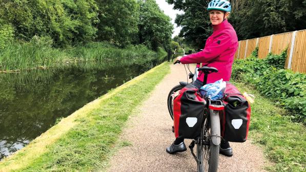 A woman sat on a bicycle laden with full rear panniers turns to look at the camera. She has paused on a canal towpath