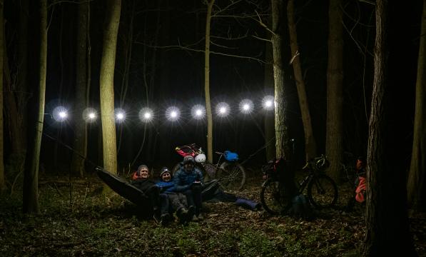 Three men sit in a hammock in some woods at night lit up by a string of fairy lights