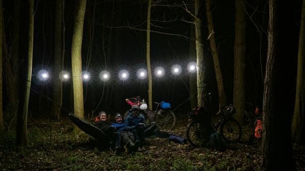 Three men sit in a hammock in some woods at night lit up by a string of fairy lights
