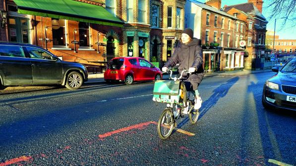 A woman cycles along a main road lined with shops and parked cars. She is wearing casual winter clothes
