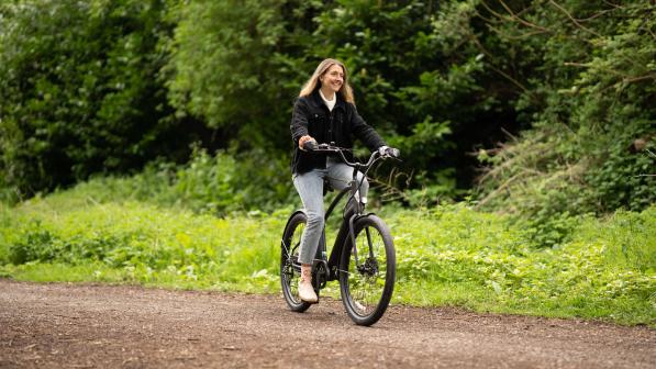 Woman wearing black jacket and blue jeans smiles as she pedals black e-bike through park, trees and grass in background.