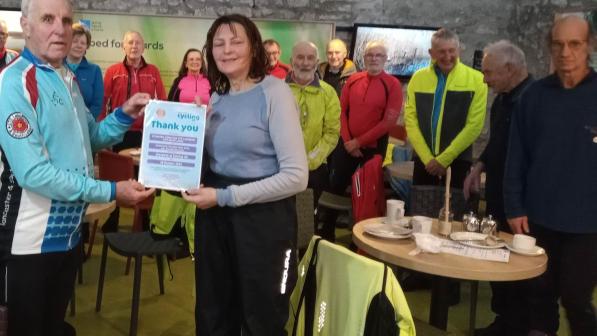 A man and woman pose for the camera holding a certificate of thanks, a group of cyclists stand behind in a cafe