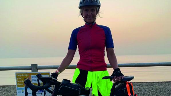 A woman is standing against a metal fence with the sea in the background. She's holding a road bike and wearing cycling kit.