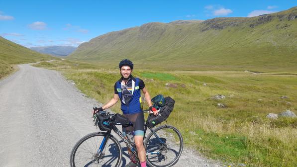 A man stands on a gravel track between mountains. His bicycle is loaded down with bikepacking bags